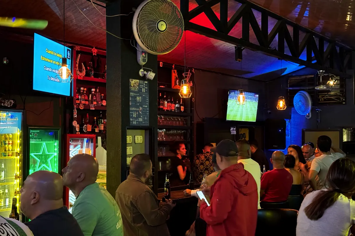 La barra llena de gente, con el karaoke en una pantalla y un partido de fútbol en la otra.
