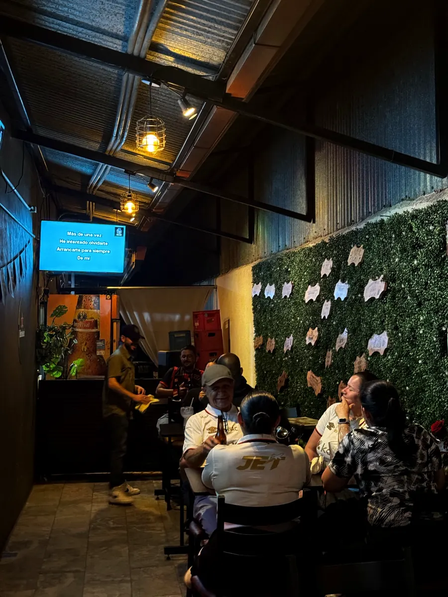 El salón de noche, con la pantalla del karaoke encendida y las mesas ocupadas junto a la pared de plantas.